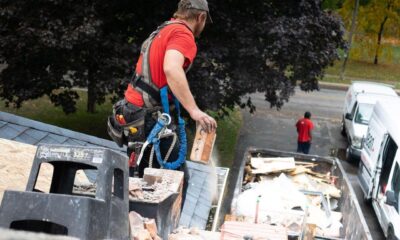 A roofing contractor wearing a safety harness removes damaged brick from a deteriorating chimney on a residential roof, with a debris bin and service vans parked on the street below.