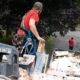 A roofing contractor wearing a safety harness removes damaged brick from a deteriorating chimney on a residential roof, with a debris bin and service vans parked on the street below.