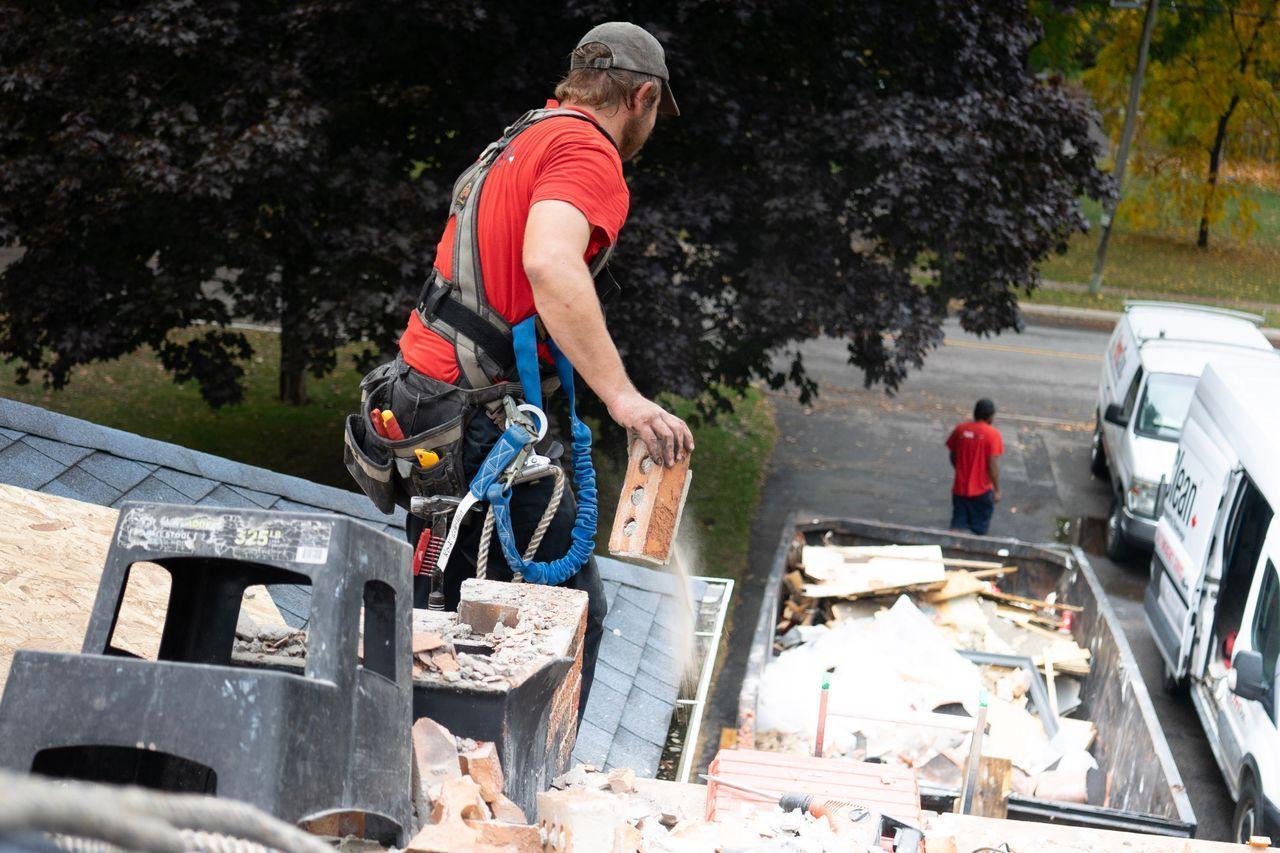 A roofing contractor wearing a safety harness removes damaged brick from a deteriorating chimney on a residential roof, with a debris bin and service vans parked on the street below.